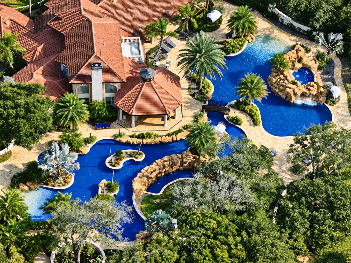 Aerial view of an estate with a winding lagoon pool, rock waterfall grotto, and arched bridge